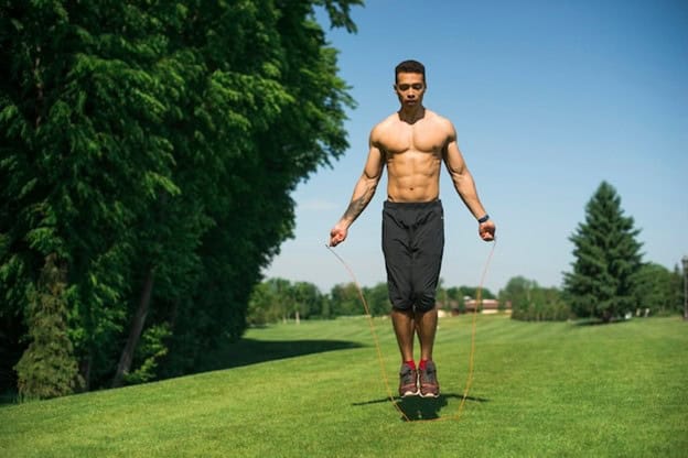 A muscular man skipping rope on a lush green field under a clear blue sky