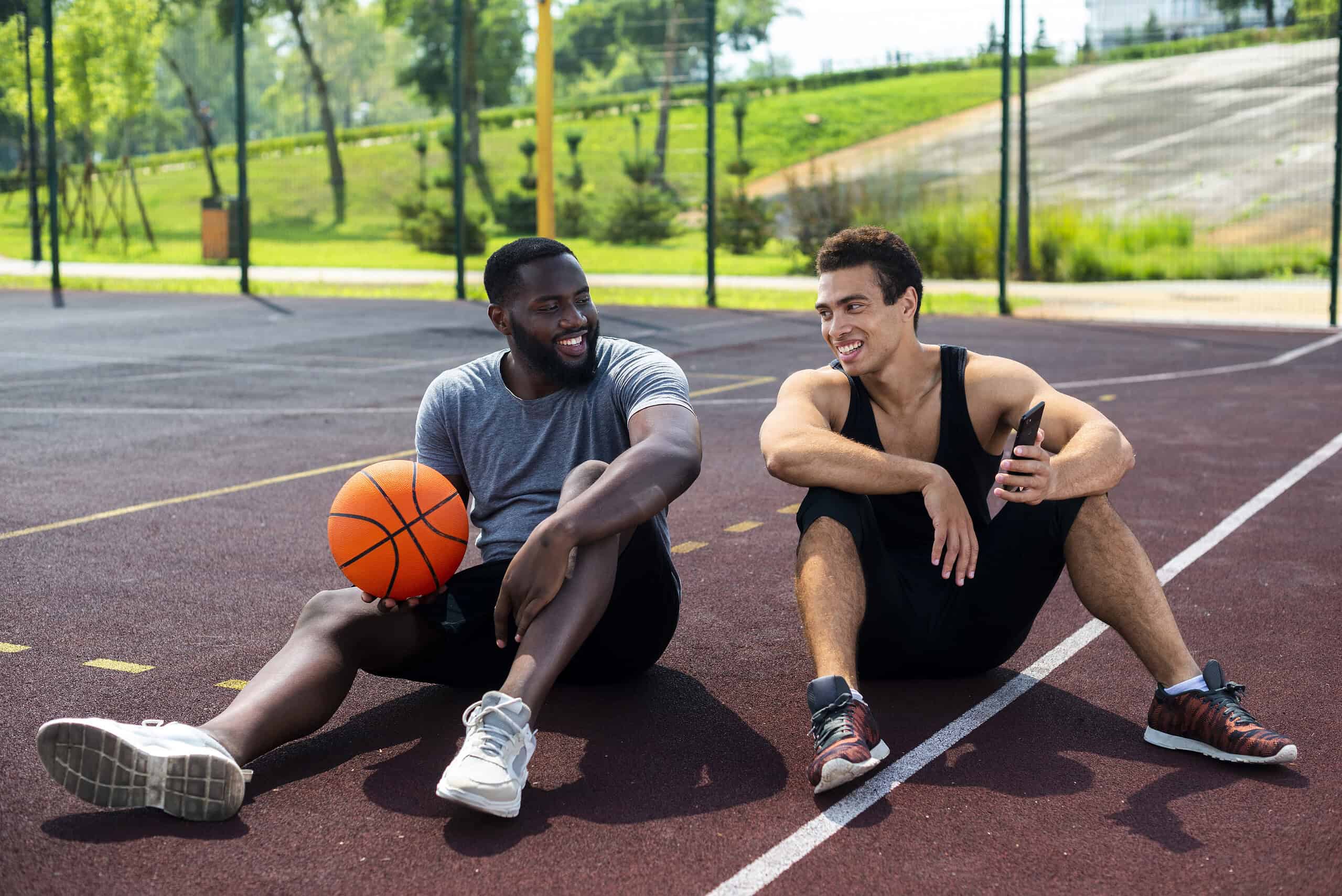Two hybrid athletes sitting on a basketball court, engaging in a casual conversation post-training