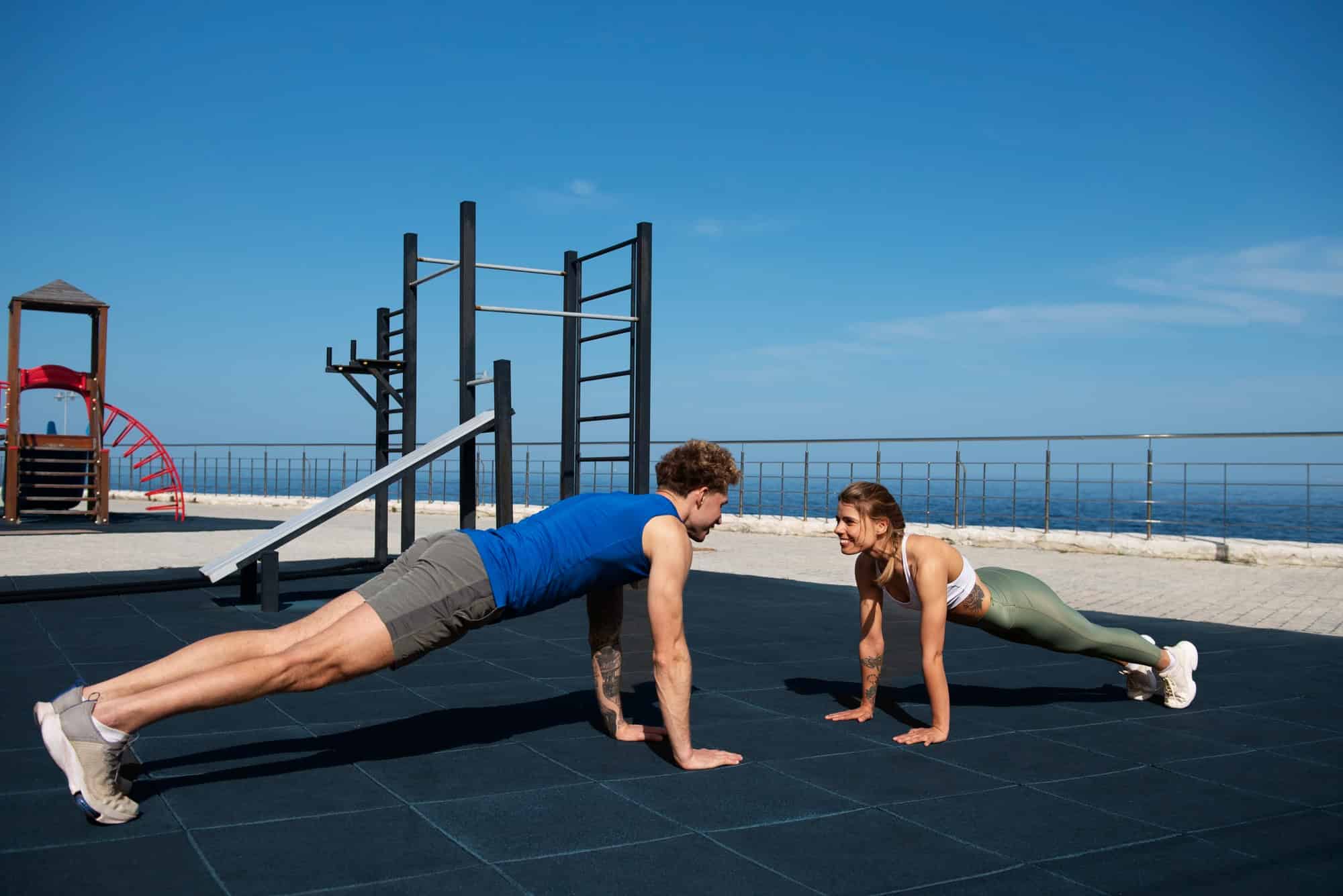 Two athletes performing push-ups on an outdoor fitness playground by the sea, demonstrating a calisthenics workout with bodyweight exercises.